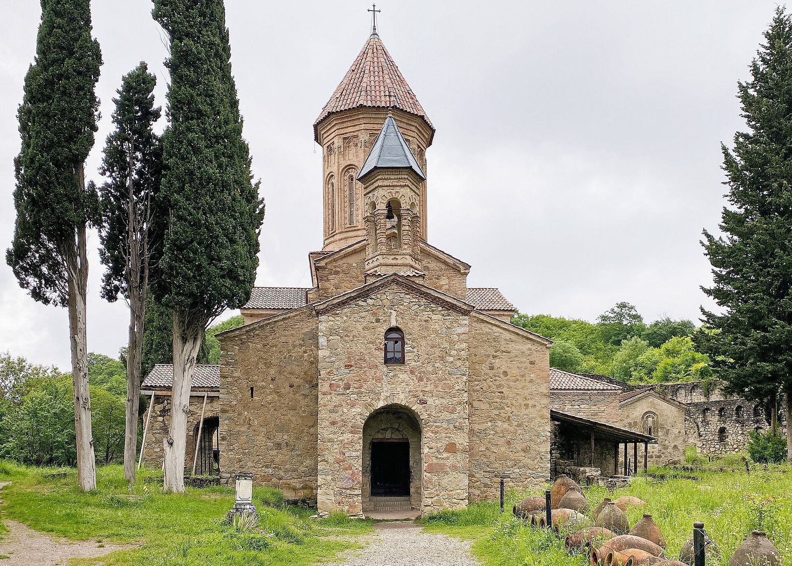 The stone Church of the Transfiguration at Ikalto Monastery between tall cypresses, with old qvevri lying on the grass