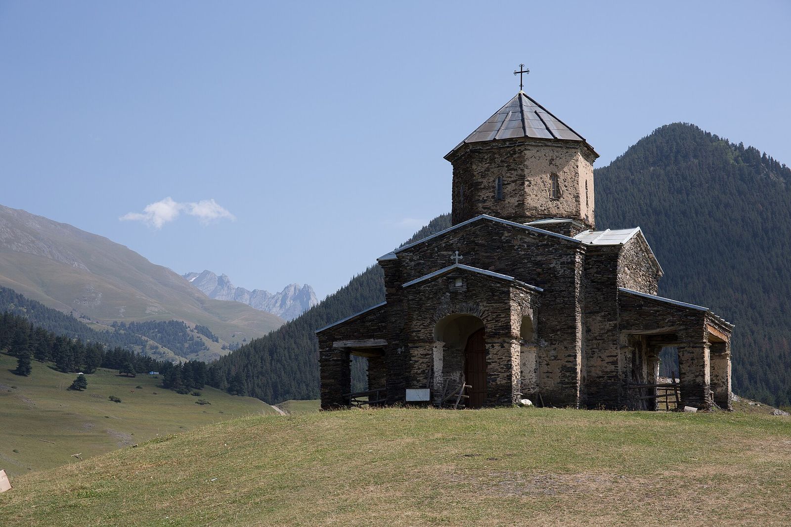 The stone church of St George in Shenako on a grassy hill, with pine forest and rocky peaks behind
