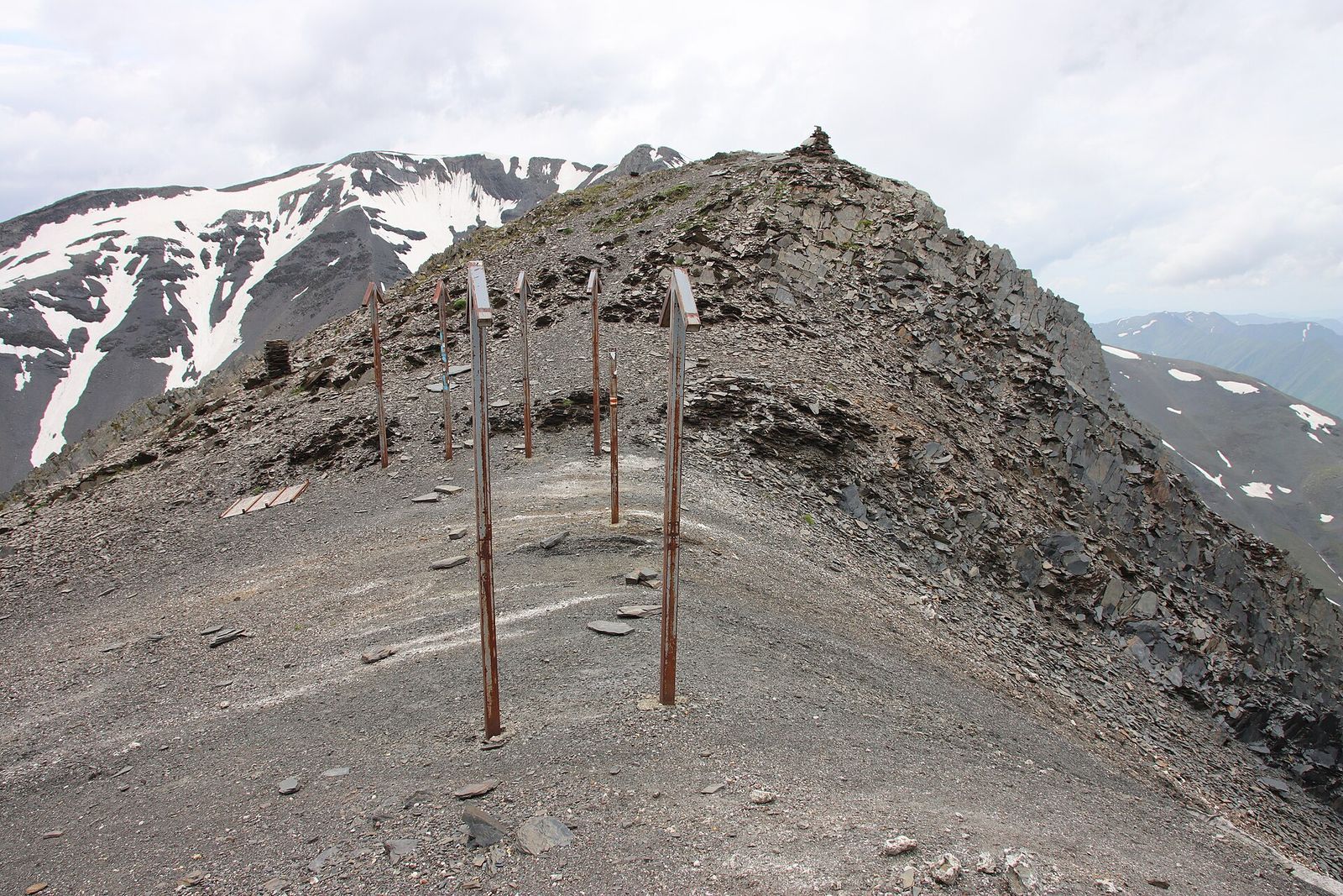 Rusted iron marker posts on the bare, rocky saddle of the Atsunta Pass, with snow-streaked peaks behind