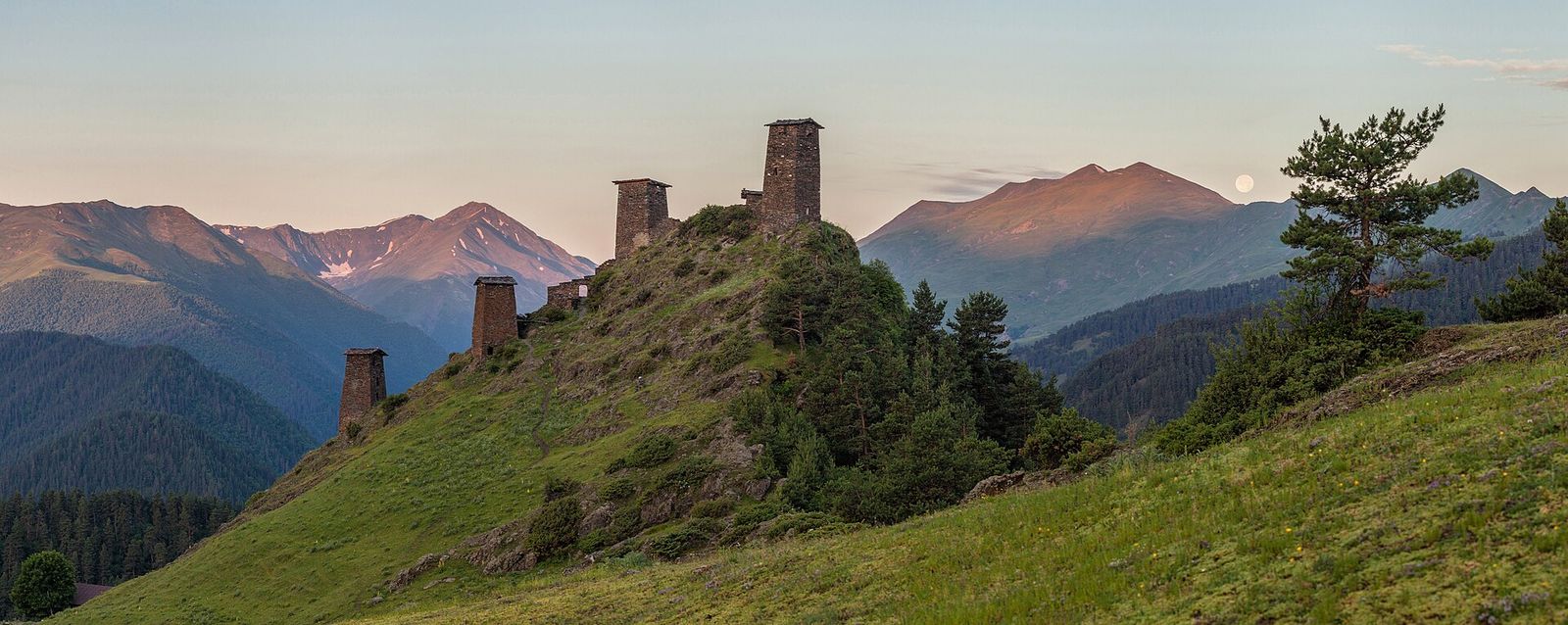 The slate defense towers of Keselo fortress on a grassy ridge above Omalo at dawn, with Caucasus peaks behind