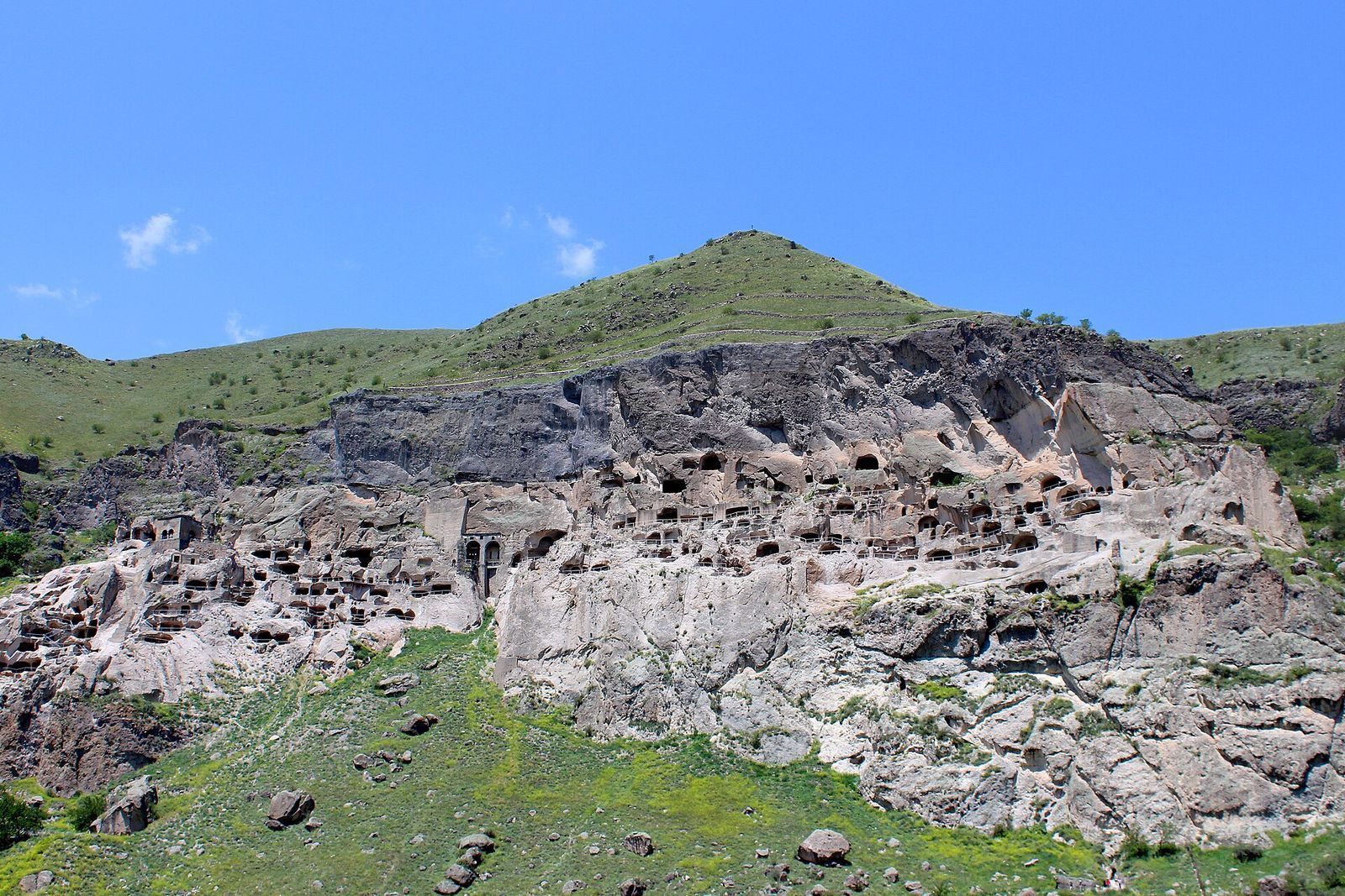 The Vardzia cave city seen from the valley, rows of cave openings across the cliff face under a green hill