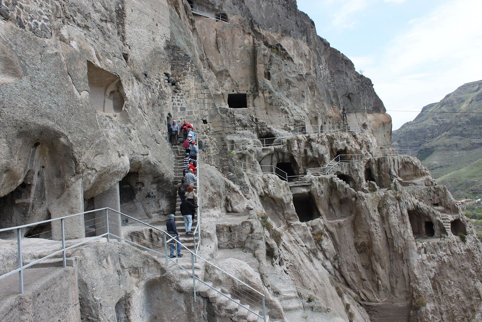Visitors climbing a stone stairway past rows of cave cells cut into the cliff at Vardzia