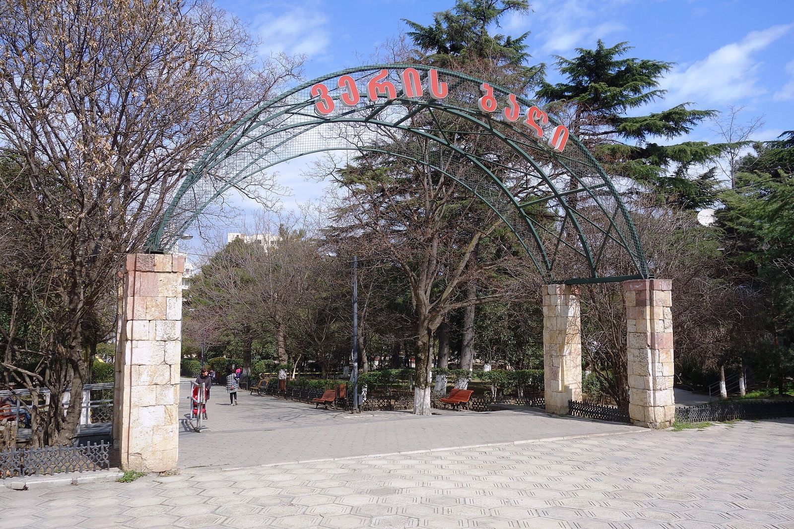 Green metal arch with red Georgian lettering over the entrance to Vera Park, with stone pillars, benches and cedar trees behind