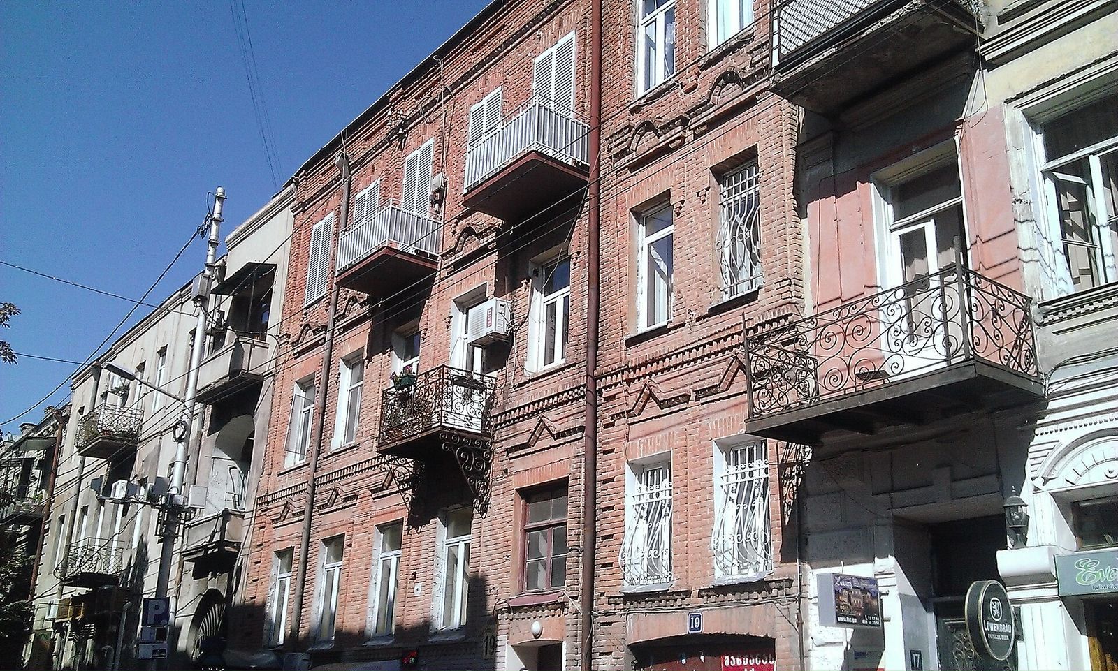 Red-brick house at 19 Giorgi Akhvlediani Street with white shutters and wrought-iron balconies, and a bar sign beside the door of number 17