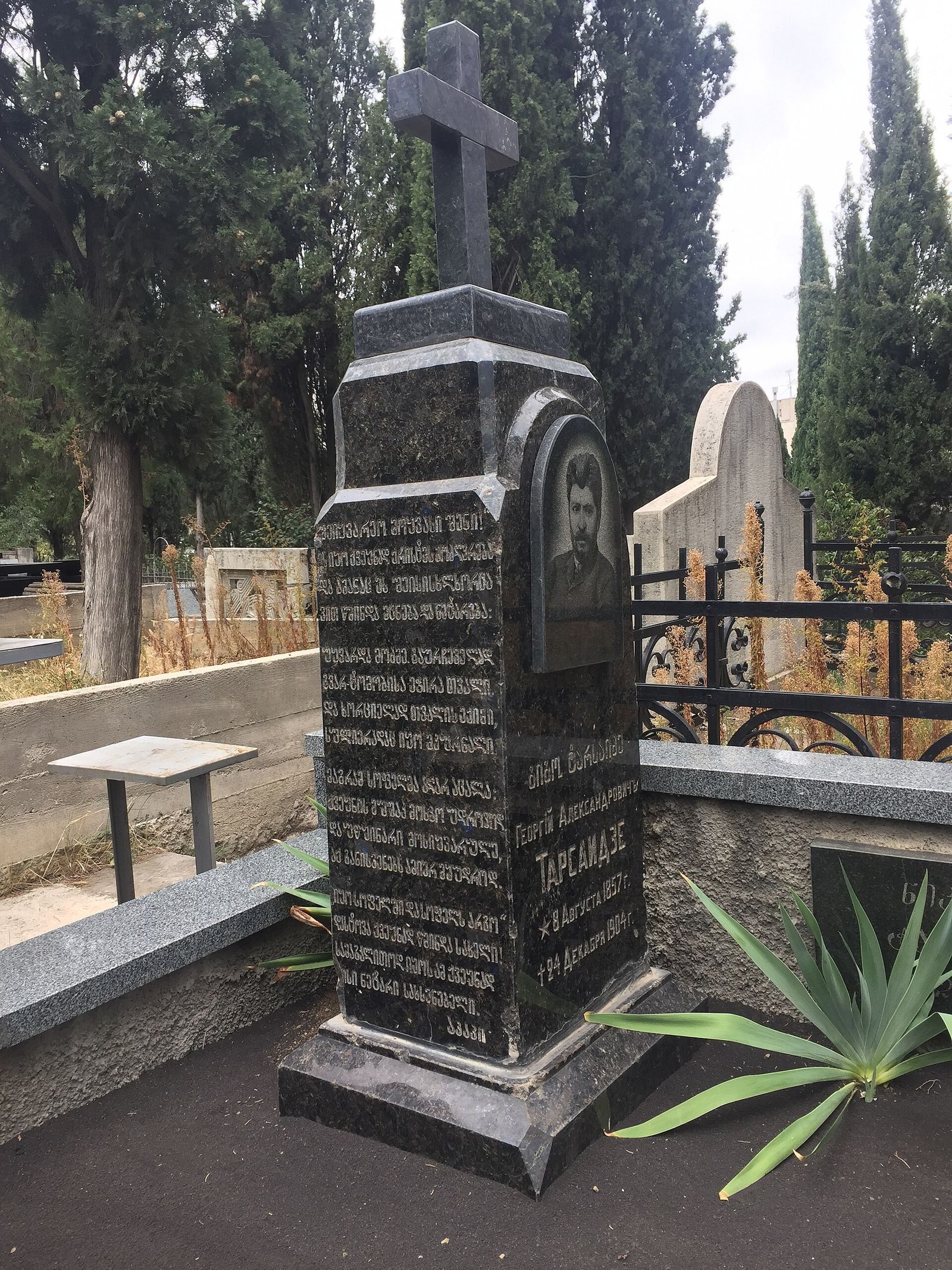 Black granite gravestone of Giorgi Tarsaidze, who died in 1904, topped with a cross among cypress trees in Vera Cemetery