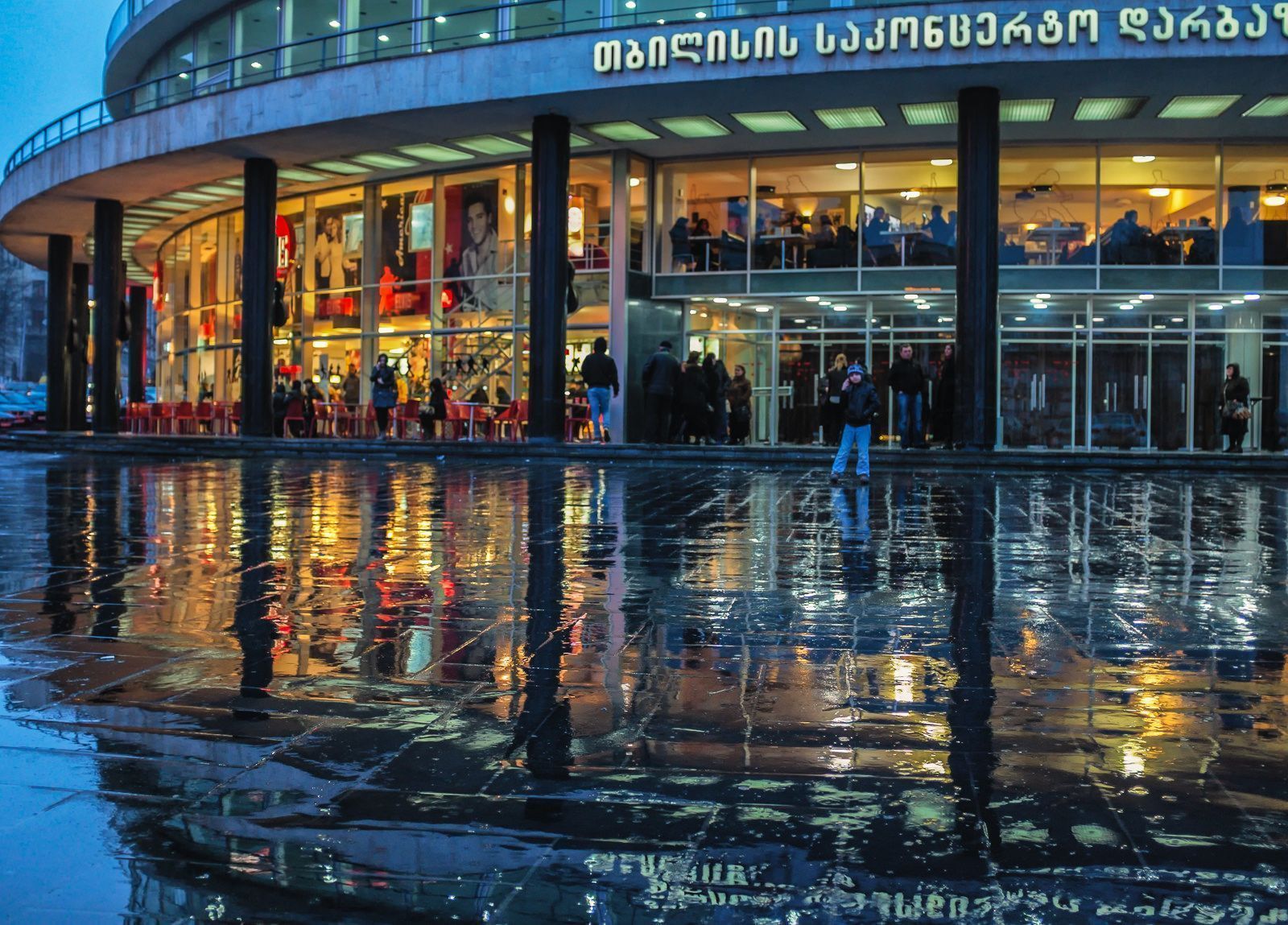 Curved glass front of the Tbilisi Concert Hall at dusk with Georgian lettering on the facade, reflected in the wet square