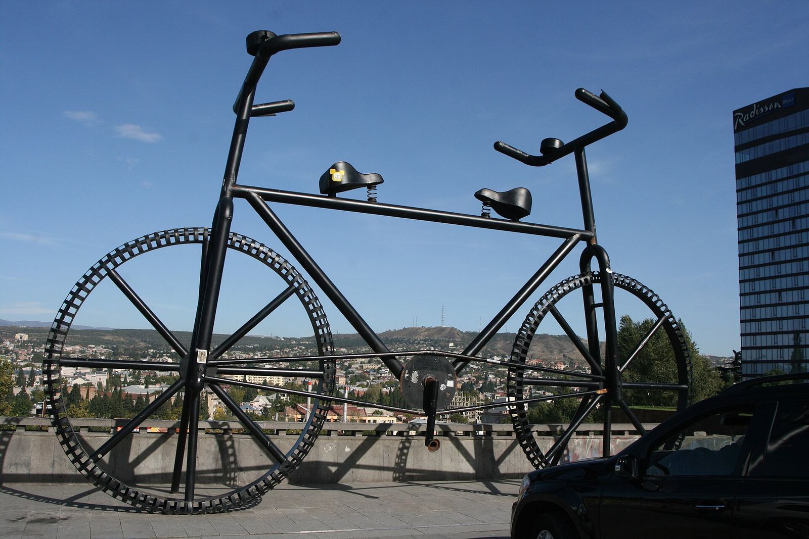 Giant black steel bicycle sculpture on Rose Revolution Square, with the Radisson Blu tower on the right and the hills of Tbilisi behind