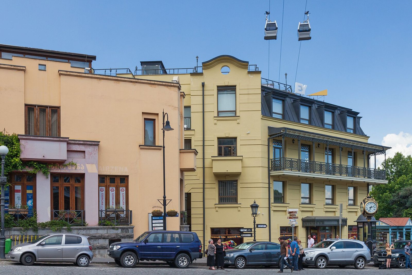 Yellow and peach facades on Kote Afkhazi Street, with an old street clock and cable car cabins passing overhead