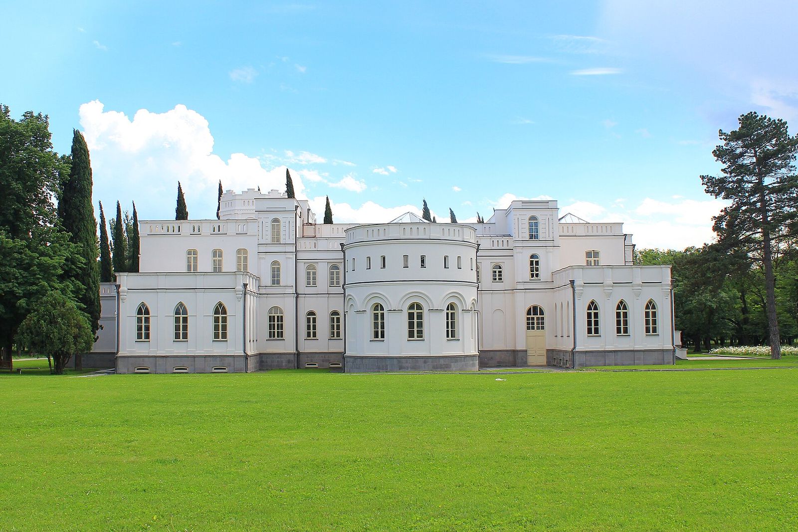 Chateau Mukhrani's restored palace seen across the wide lawn of its gardens, framed by cypresses and pines
