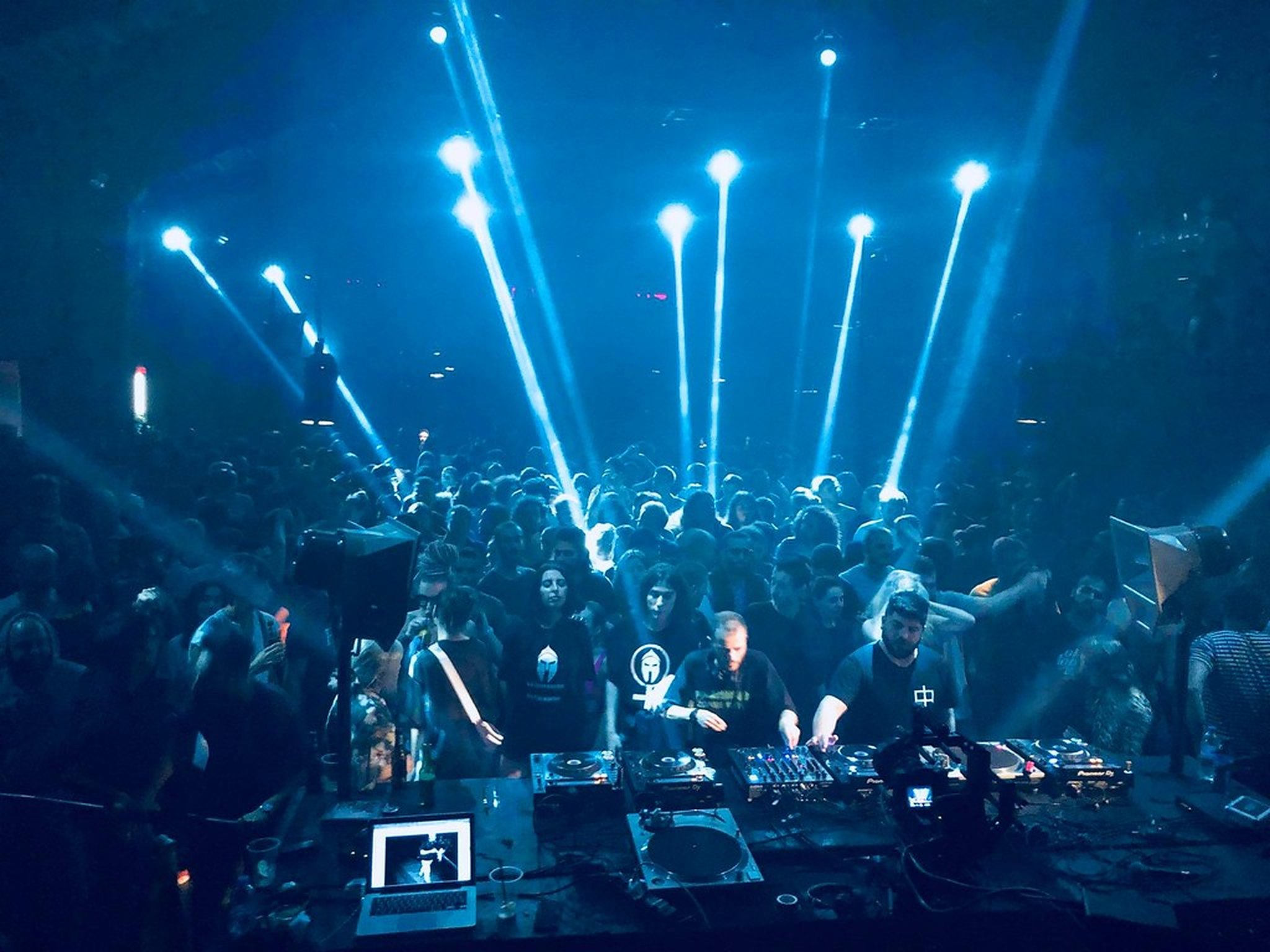 DJ at the decks facing a packed crowd under blue light beams